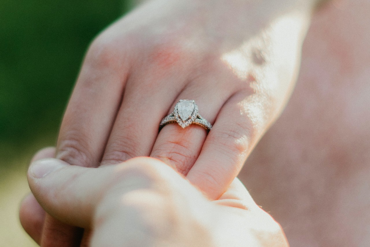 A man’s hand holding a woman’s that is adorned with a pear shape halo engagement ring.