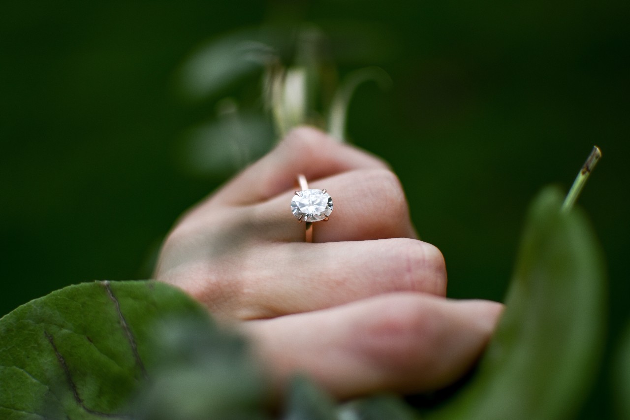 A woman’s hand, wearing an oval engagement ring, holding a bouquet of greenery.