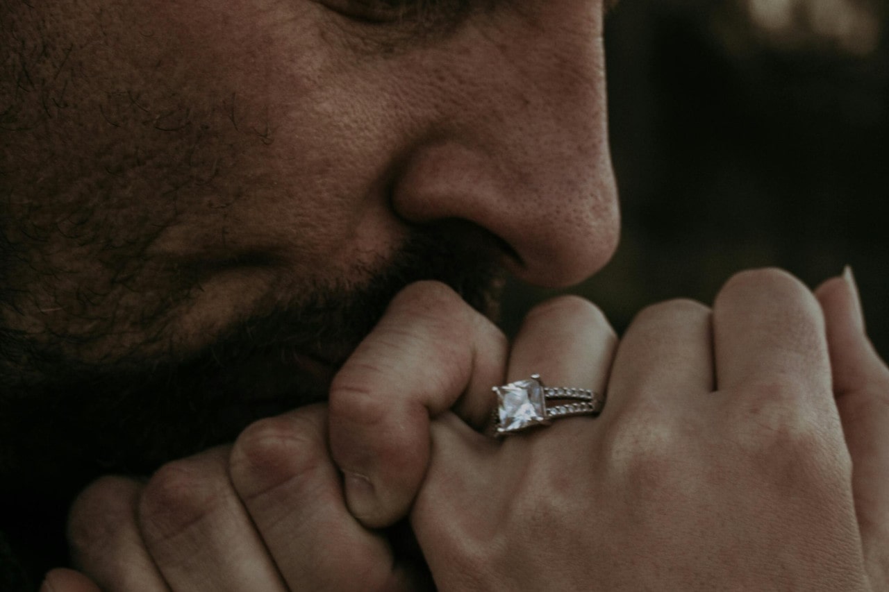 a man kissing a woman’s hand that is adorned with a princess cut engagement ring