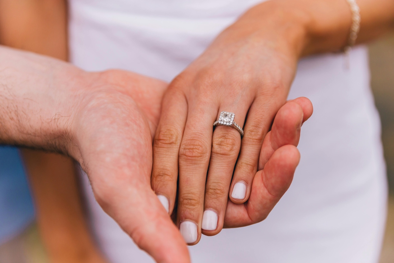 a man’s hand holding a woman’s that is adorned with a princess cut halo engagement ring