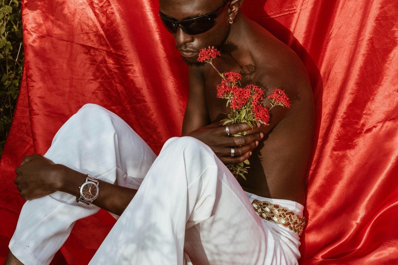 a man sitting in front of a red silk sheet, wearing a silver watch and silver jewelry