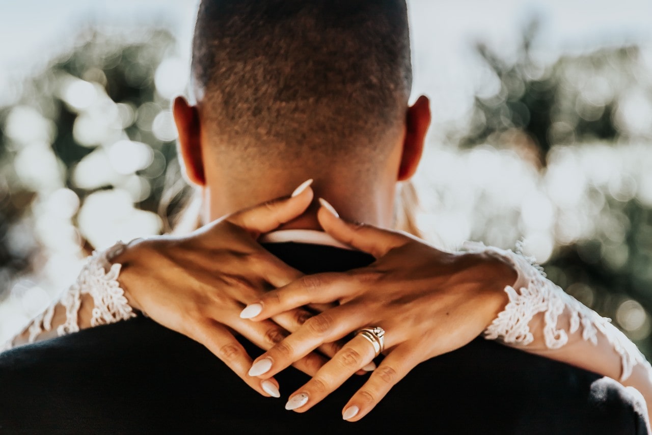 a bride's hands clasped behind the neck of her groom–wearing a white gold engagement ring and wedding band