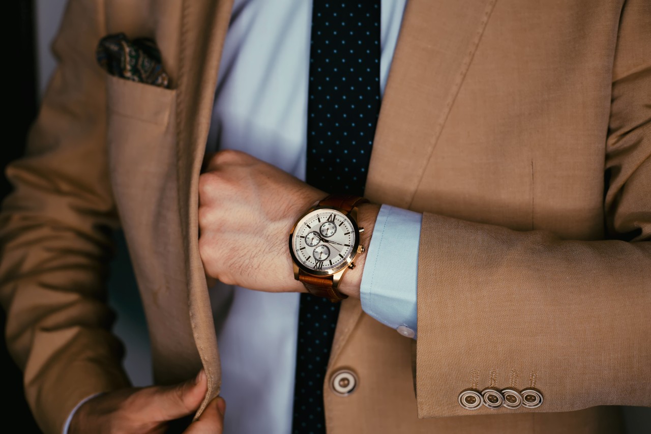 A man wearing an impressive yellow gold and brown leather watch reaching into his tan suit jacket.