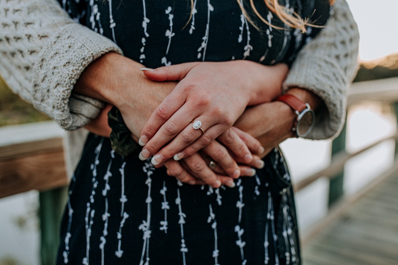 A couple hugging on a boardwalk; the woman has a pretty yellow gold diamond halo ring and the man person has a wedding band and watch.